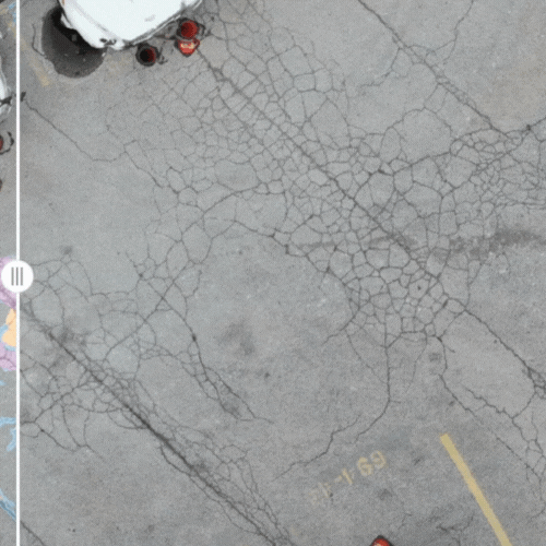 Aerial view of a large, cracked concrete surface with yellow parking lines. Two white vehicles and people in red outfits positioned near the top and bottom edges. Cracks create irregular patterns across the concrete.