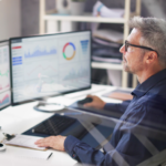 A man with gray hair and glasses is sitting at a desk with three computer monitors displaying various graphs and charts. He is holding a pen, and there is a notebook on the desk. Shelves with books are in the background.