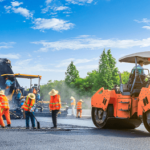 Construction workers in orange vests and hats operate heavy machinery while paving a road on a clear day. A steamroller is prominent on the right, with green trees and a blue sky in the background.