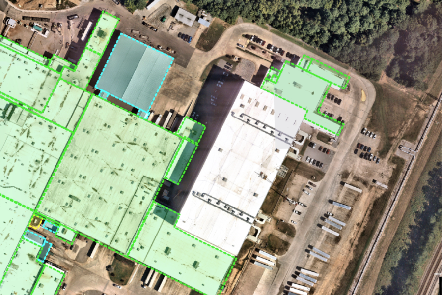 Aerial view of an industrial facility with large, white-roofed buildings outlined in green—ideal for commercial real estate property owners and facility managers to prepare for and respond to hurricane season—surrounded by lots, vehicles, and trees.